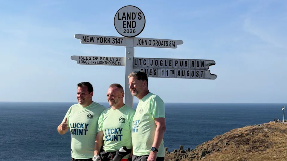 Gary McKee standing at Land's End with two members of his running support team. All are wearing mint green T-shirts with the words "lucky saint" on the front. They are standing next to a black and white finger signpost detailing distances from Land's End to other places, such as New York and John O'Groats. The men are pictured side-on posing for a picture. They are all smiling and one is giving the camera a thumbs up.