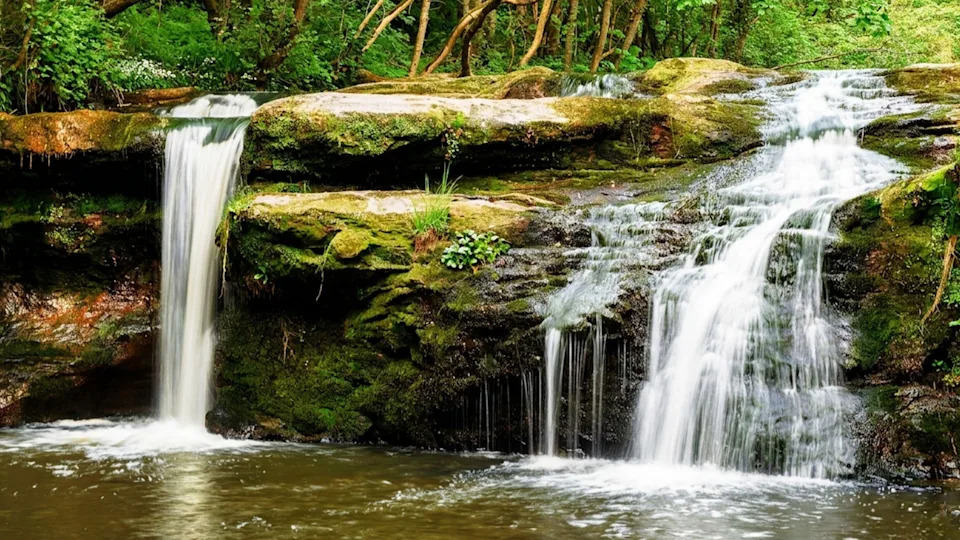 A woodland waterfall cascades over moss-covered rock ledges into a calm pool below.