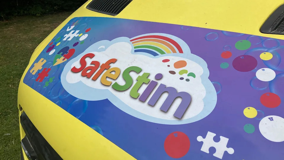 A sign of the Safestim logo which is on front of the sensory van. It has a rainbow font which is in a cloud with a rainbow above on the background of a purple and blue background. There are other symbols around the logo which includes bubbles and puzzle shapes.