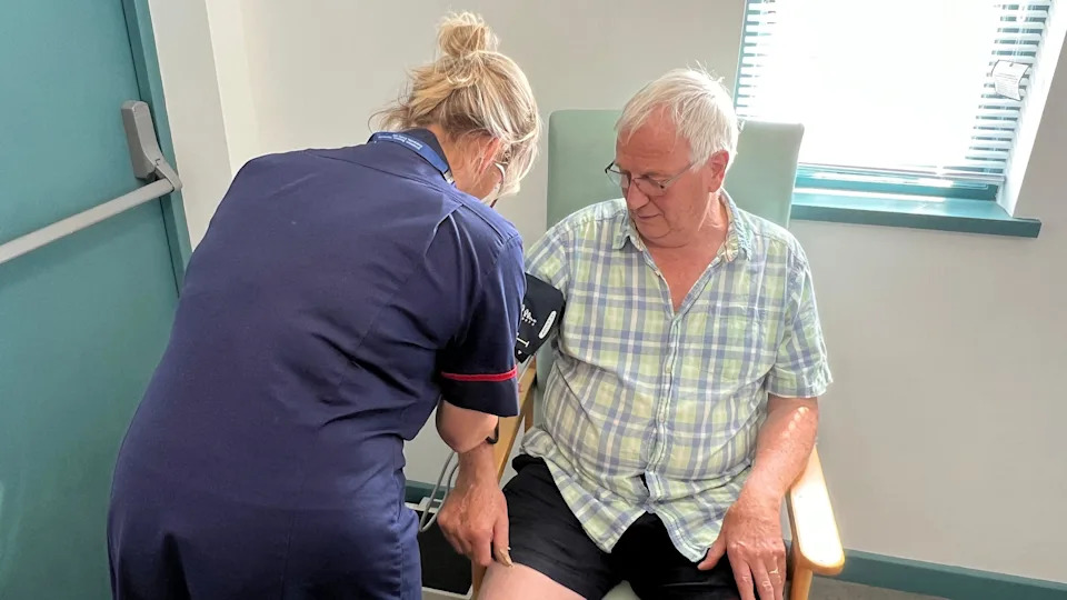A man with grey hair wearing glasses and a blue and green checked shirt is having his blood pressure taken by a nurse.