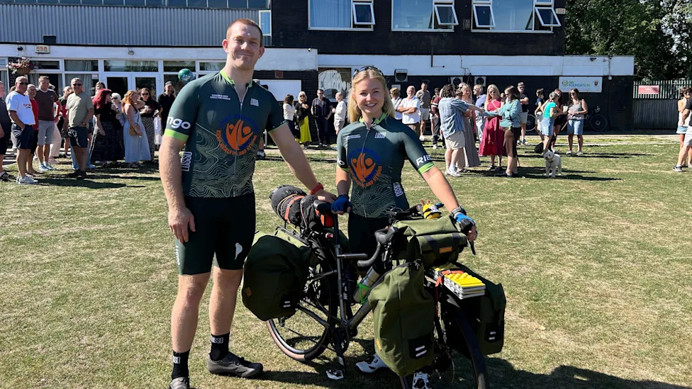 Lucy and Ciaran stand together with a bike that is loaded with panniers, wearing matching, green cycling jerseys. It is a sunny day and they are standing on grass, with a crowd of people mingling in the background.