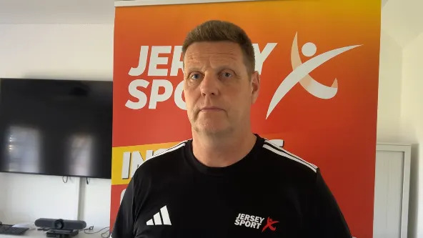 A man is pictured in an office. He is wearing a Jersey Sport branded tshirt. He has short brown hair and has a serious look on his face. In the background are some posters for Jersey Sport.