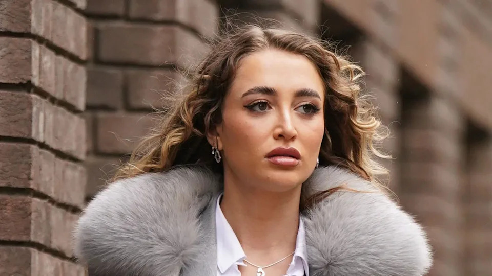 Georgia Harrison with long curled blonde hair, looking slightly away from the camera with a brick wall behind her. She is wearing a grey jacket and white shirt, and her overcoat has a big, fluffy grey collar.