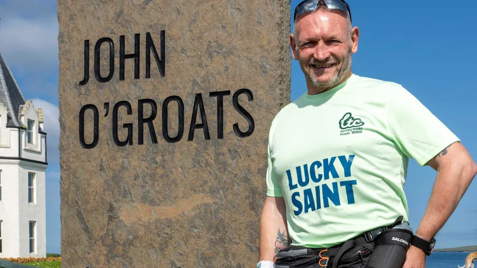 Gary McKee at the start line of his 900-mile challenge in John O'Groats. He is standing next to a stone feature with an inscription of John O'Groats. He has short salt and pepper hair and short beard. He is smiling at the camera and is wearing sunglasses perched on top of his head and a mint green T-shirt with a logo.