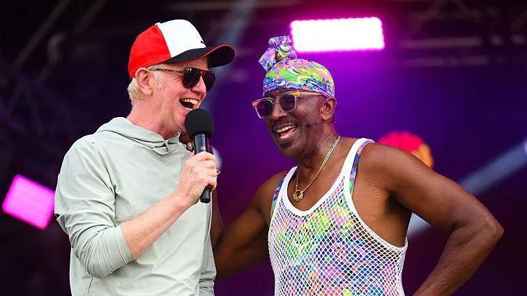 Chris Evans and Mr Motivator on stage at CarFest. Evans is wearing a red and white baseball cap, dark glasses and a light-coloured cotton hoody. Mr Motivator is wearing a colourful scarf on his head, dark glasses and a mesh vest over a colourful vest. Both men are laughing.