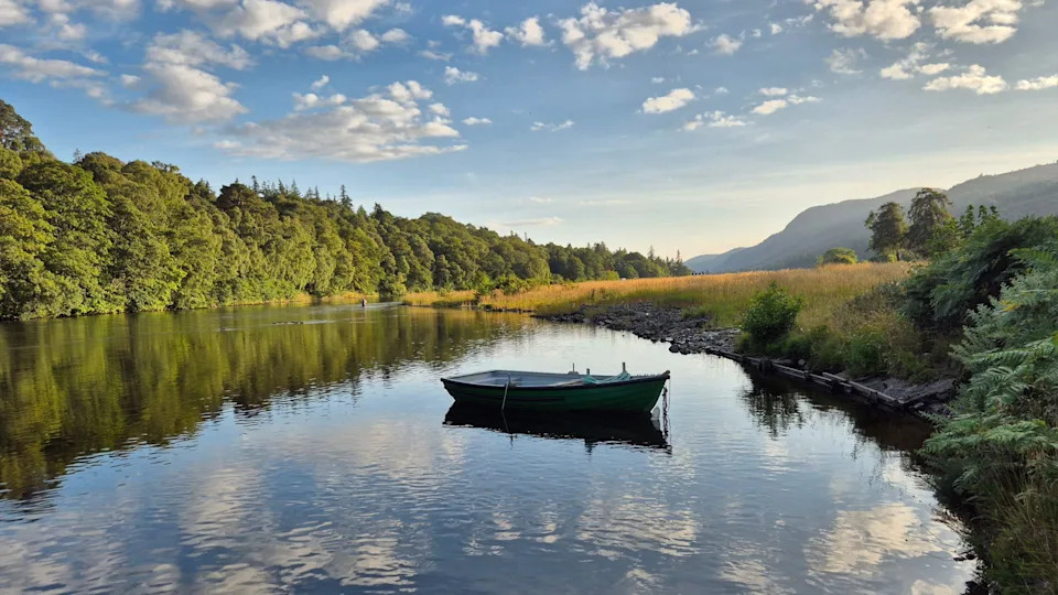 A small green rowing boat floats on calm water beside the shore of Loch Ness.