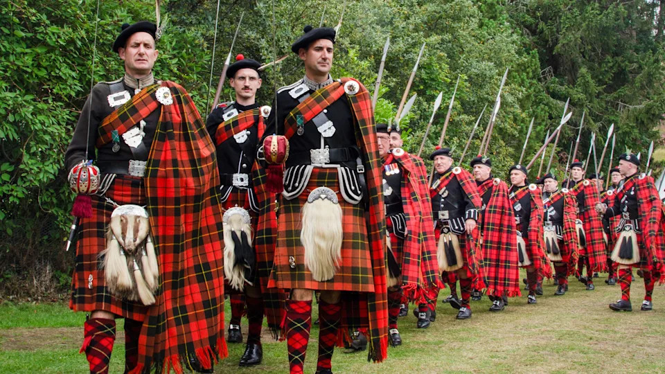 A procession of Lonach Highlanders in traditional tartan dress marches across a grassy field, carrying long pikes and wearing matching red and black tartan plaids.
