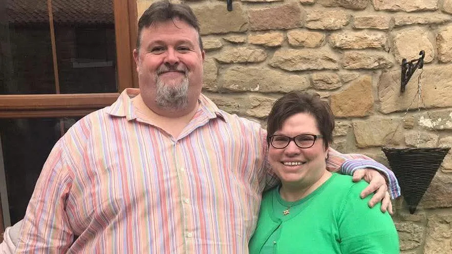 Steffen and Becky Peddie stand in front of a stone cottage. Steffen, on the left, is wearing a striped pink shirt, has a grey beard and dark hair. He is of a large build. He has his arm around his wife who is quite a lot shorter than him, she also has dark hair, is wearing glasses and has a green twinset on.