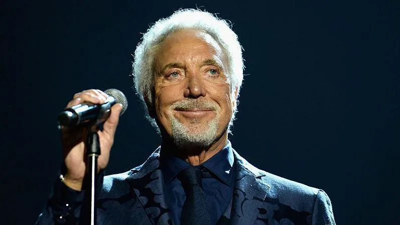 Sir Tom Jones holds a mic stand and smiles. He is wearing a dark suit over a dark shirt and tie. He has white hair and white goatee-style beard.