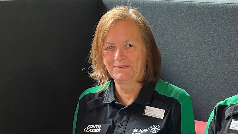 A picture of Viv Moore smiling looking at the camera wearing a StJohn's Ambulance black top with green shoulders, a Youth Leader badge.