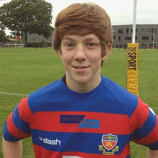 A young Ciaran Booth stands on a rugby pitch wearing a red and blue striped rugby shirt.