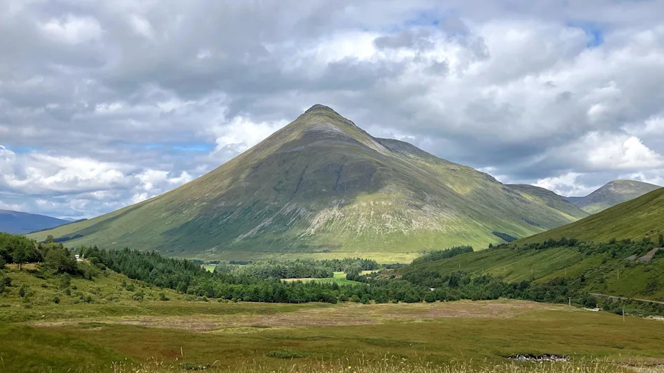 A dramatic conical mountain rises above a green valley, illuminated by patches of sunlight beneath a sky filled with large grey and white clouds.