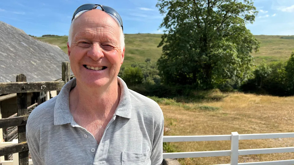 A man wearing a grey polo top with sunglasses on his head is standing on a farm in front of a white gate on a sunny day smiling at the camera.