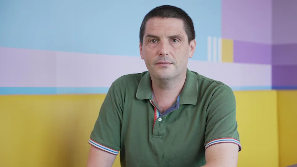 Dr Paul Evans is sitting at a table in front of a yellow seating area looks into the camera. He has dark hair and brown eyes. He's wearing a green polo shirt with blue and red piping around the sleeves.