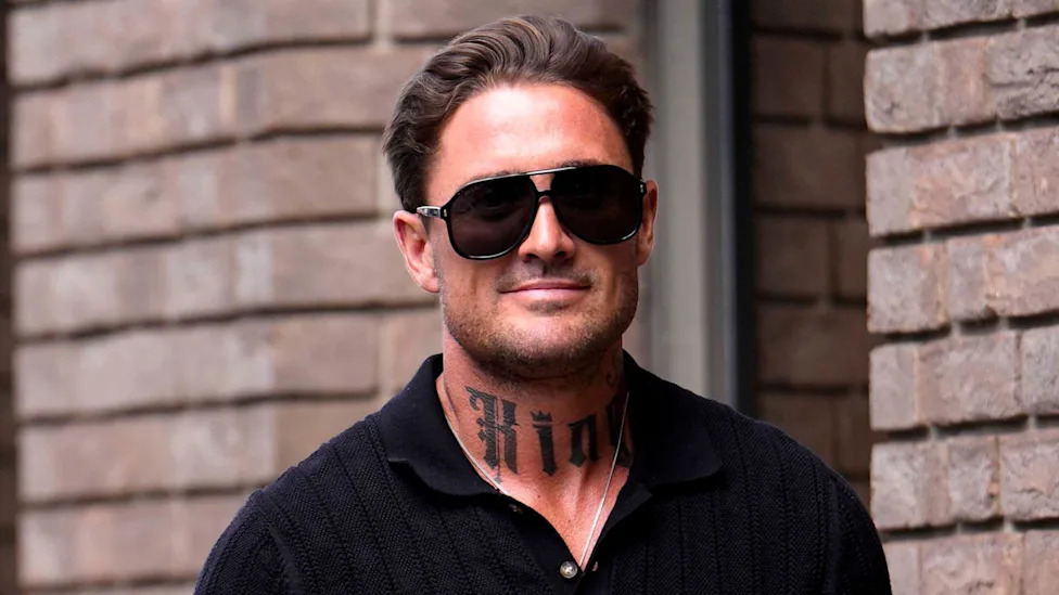 Stephen Bear smiles at the camera as he walks in to court, with a brick wall behind him. He has dark hair and stubble and is wearing big sunglasses and a black shirt. He has a prominent tattoo on his neck.