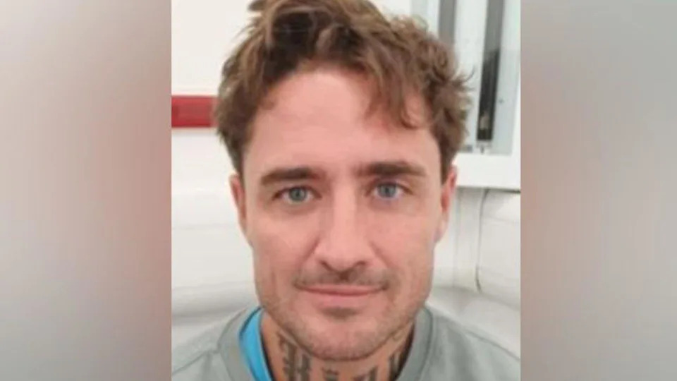 Stephen Bear custody image. He is wearing a grey jumper over a bright blue T-shirt, and he is pictured near a wall that is largely white behind him. He has tattoos on his neck and is smiling at the camera.