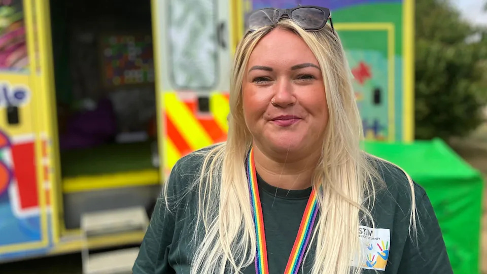 A white woman stood outside in front of an ambulance that has been transformed into a sensory van. She has blonde hair and is wearing a dark green t-shirt with a rainbow lanyard.