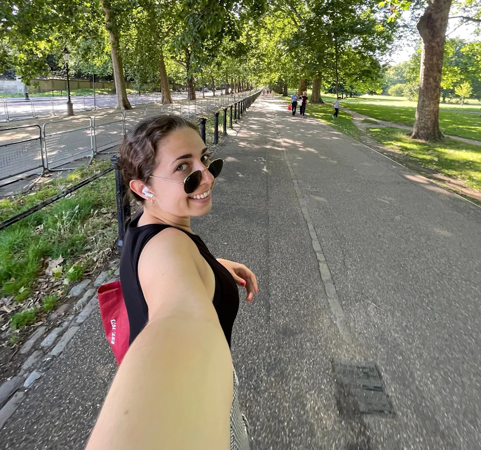 Person taking a selfie while walking in a sunny park, wearing sunglasses and earphones, smiling at the camera