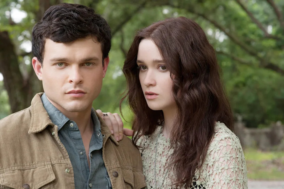 A man in a casual jacket stands with a woman in a lace top, both looking contemplative in a wooded setting