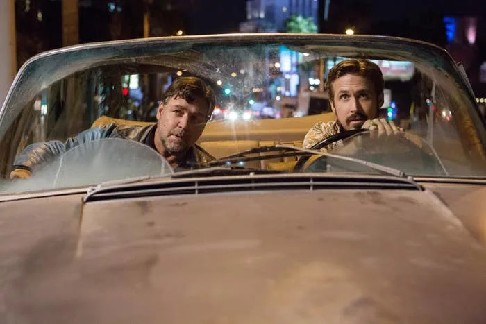 Two men sit in an old convertible, driving at night through a city. The scene suggests a classic or action movie setting