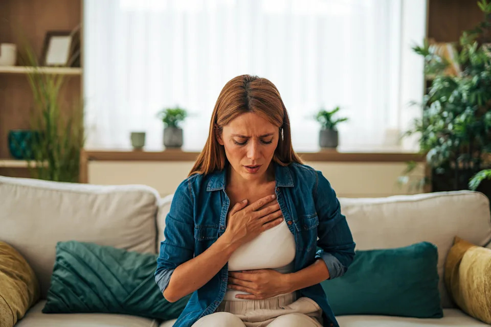Person sitting on a couch, pressing hand to chest with a concerned expression, possibly experiencing discomfort or pain