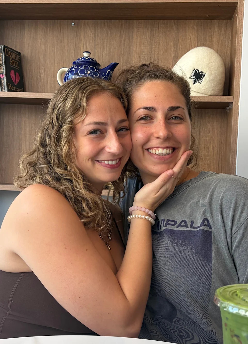 Two people smiling at a café table; one wears a sleeveless top, the other a graphic T-shirt. A drink with fruit is on the table. Books are in the background
