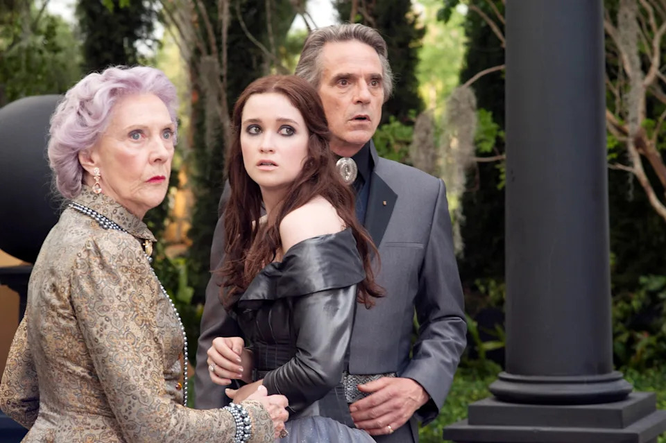 Three actors in period attire look startled, in a garden setting. The young woman wears an off-shoulder gown, the older adults in regal outfits