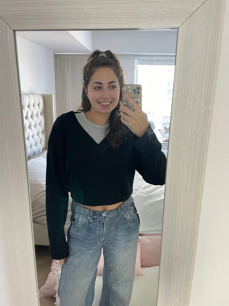 Person taking a mirror selfie in a bedroom, wearing a black V-neck sweater and light jeans, smiling at the camera