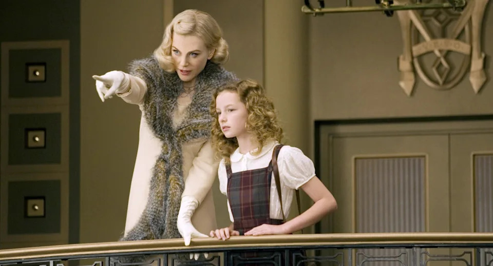 Two actors stand on a balcony. The woman wears a fur-trimmed coat, pointing ahead, while the young girl in a plaid dress looks in the same direction