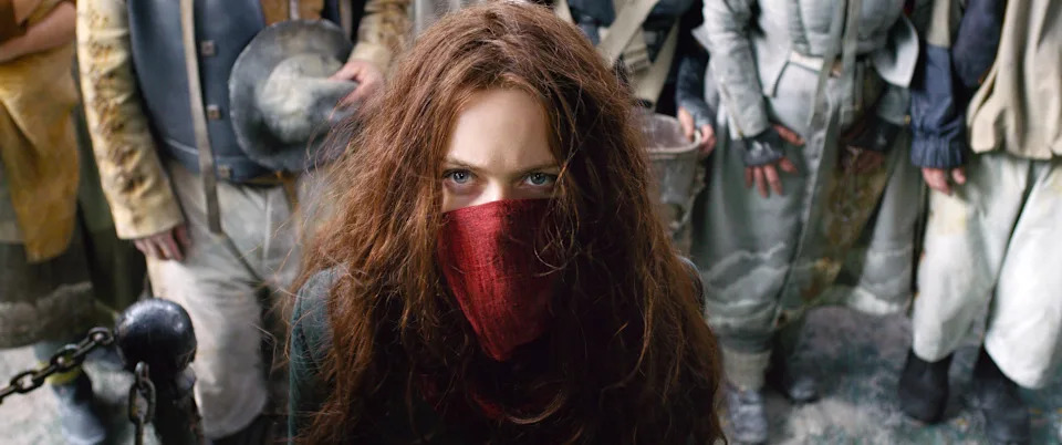 Person with long hair and a red scarf covering mouth looks directly at the camera, surrounded by people in rugged clothing