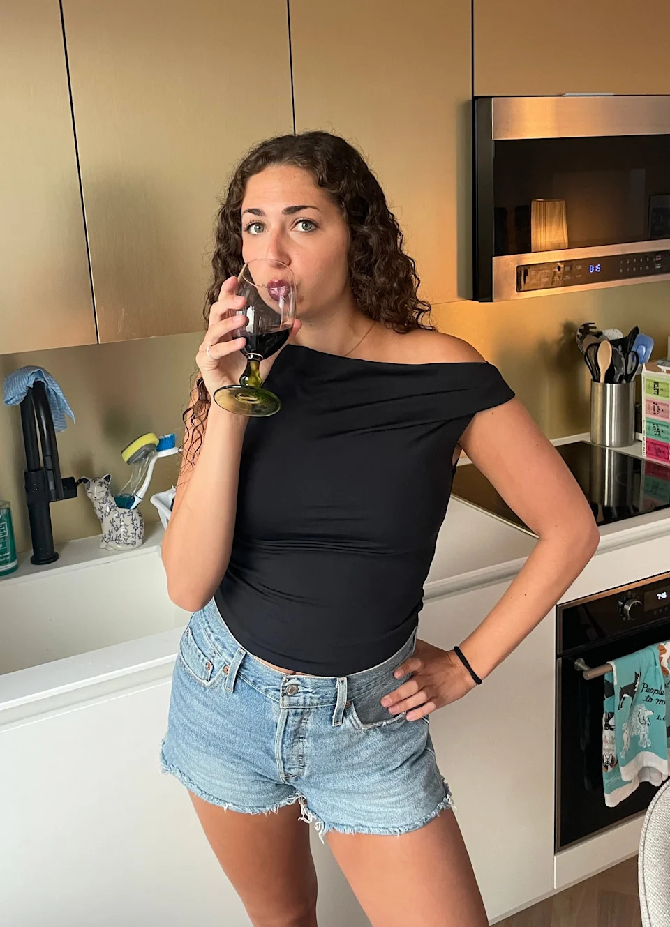Woman in an off-shoulder top and denim shorts drinks wine in a modern kitchen
