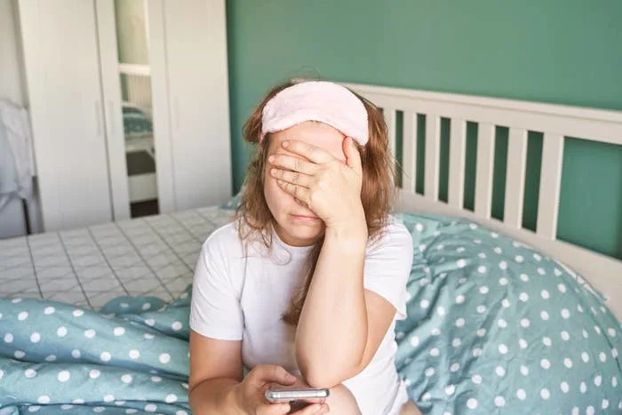 Person sitting on a bed, wearing a sleep mask on their forehead, holding a phone, and covering their face with one hand, suggesting tiredness