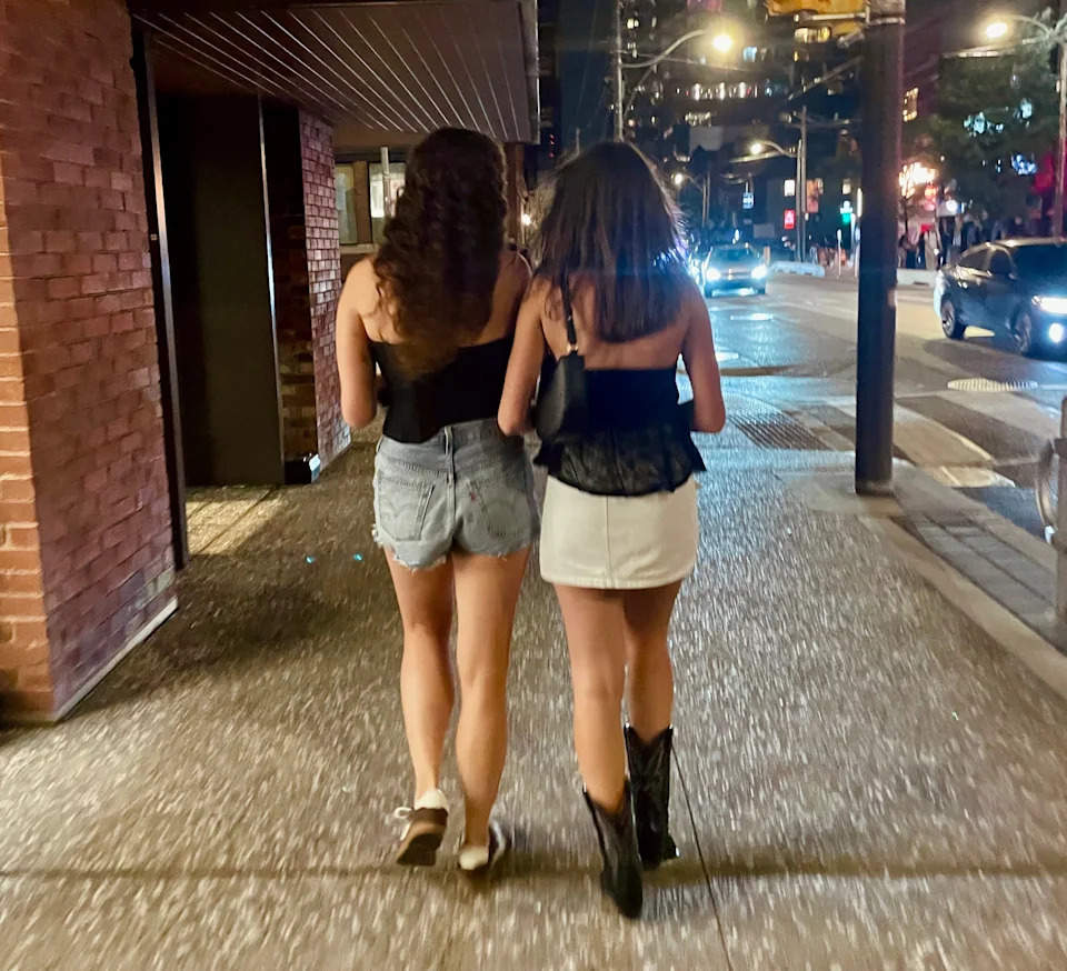 Two people walk on a city street at night. One wears denim shorts and a top, the other a short skirt and top with boots. Urban nightlife scene