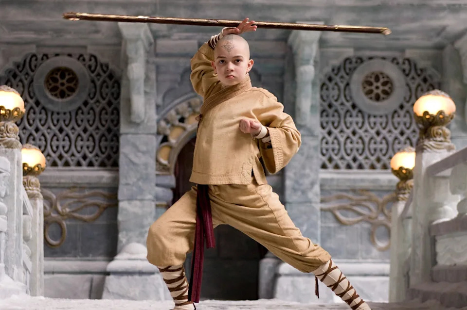 Person in a warrior pose, holding a staff above their head, in a stylized stone setting reminiscent of a fantasy series