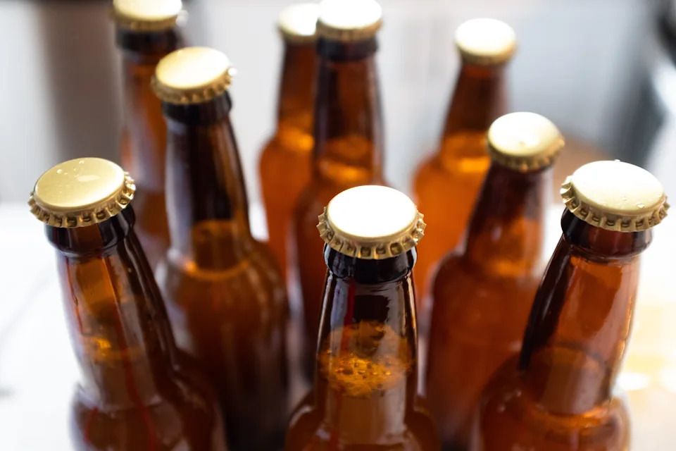 Nine brown glass beer bottles are arranged closely together, all capped and unopened, on a flat surface