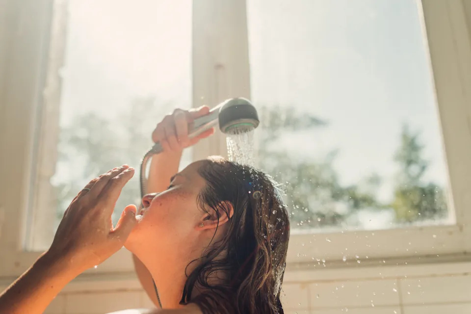 Person showering under handheld showerhead, relaxed expression, in a sunlit bathroom