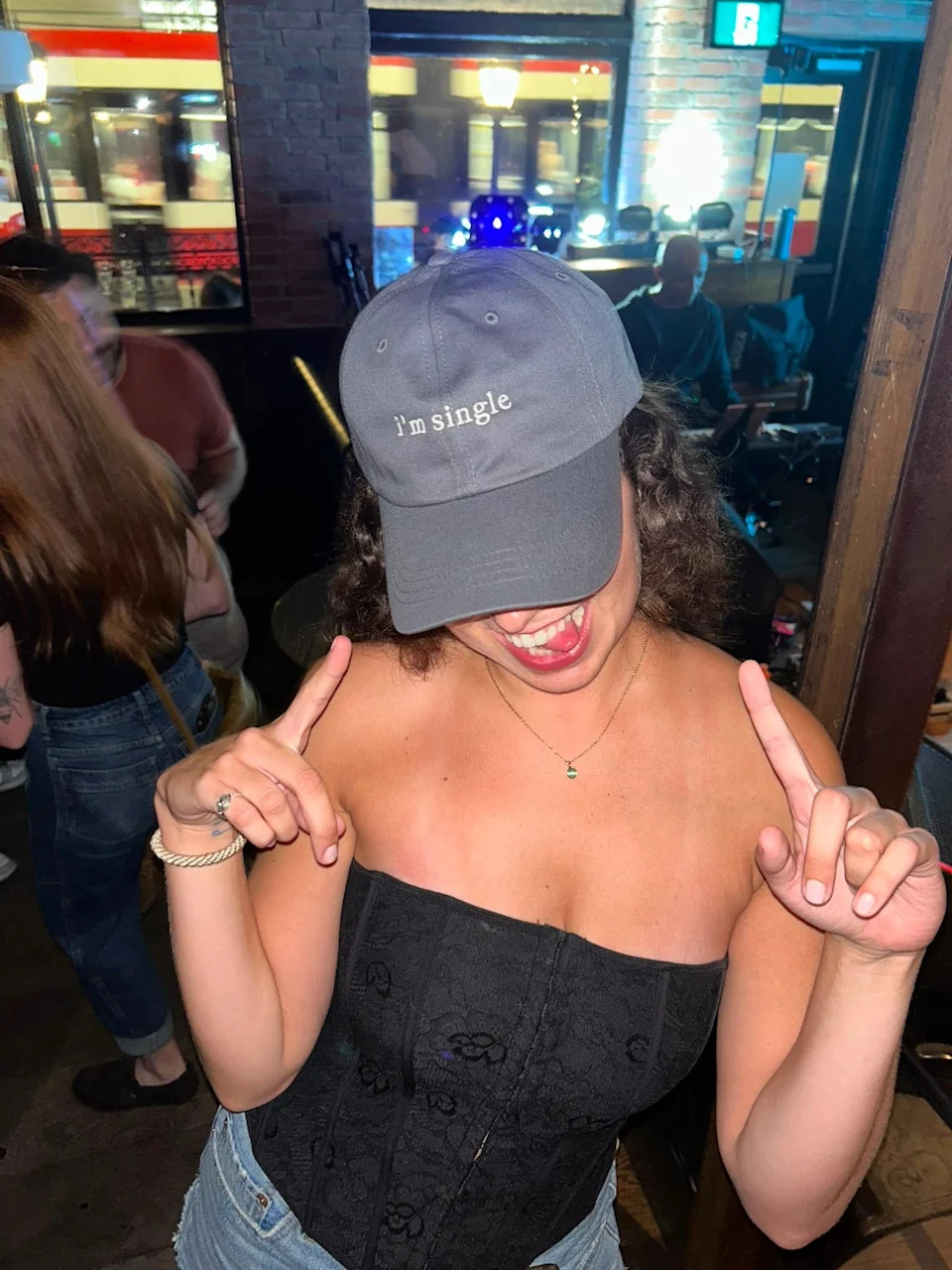 Person wearing a cap with "I'm single" text, smiling and pointing to the cap in a lively indoor setting
