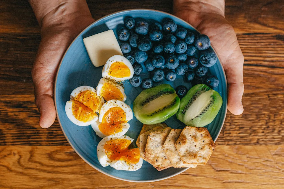 Plate with halved boiled eggs, kiwi slices, blueberries, cheese, and crackers, held over a wooden surface
