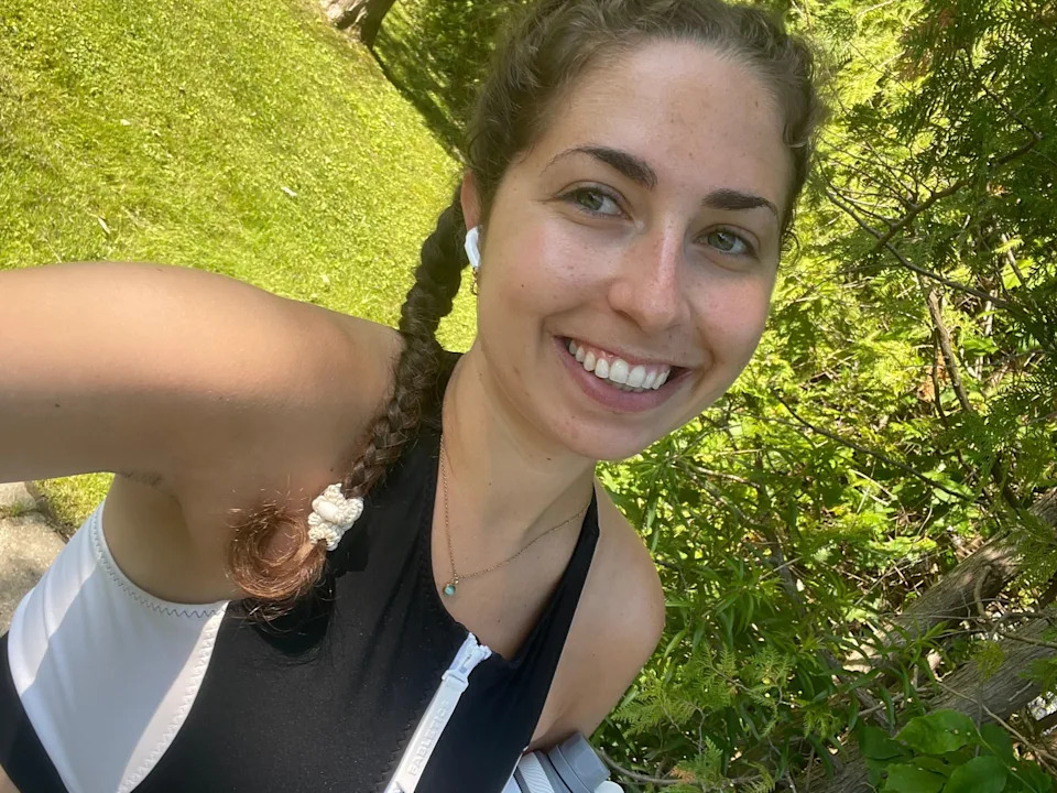 A woman with braided hair smiles outdoors, wearing a sleeveless top, surrounded by greenery