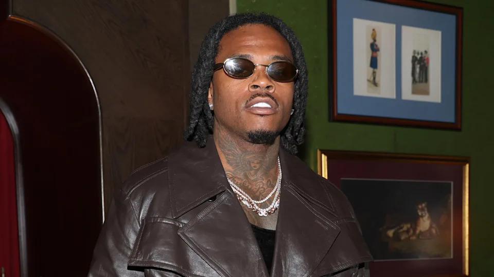 Gunna wearing sunglasses and a brown leather jacket stands in front of framed art.
