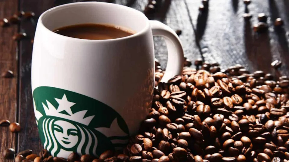 A white Starbucks mug filled with coffee sits on a wooden surface surrounded by scattered coffee beans, evoking the cozy comfort often associated with healthy foods high in sugar.