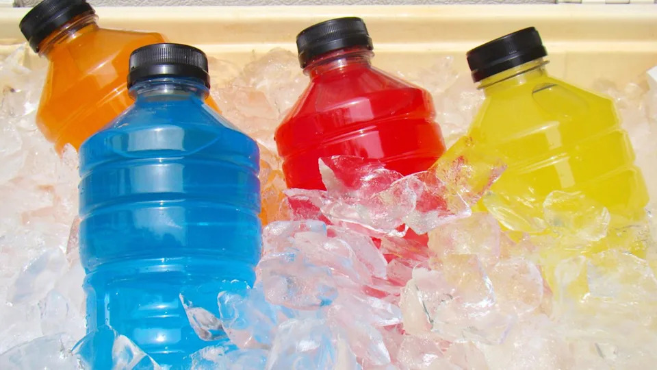 Four plastic bottles with blue, red, yellow, and orange drinks—healthy foods high in sugar—are placed on ice cubes inside a cooler. All bottles have black caps.