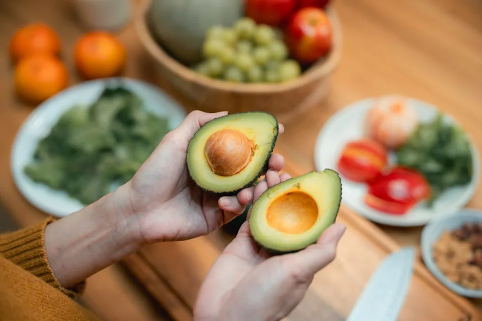 A woman's hands holding a halved avocado over a cutting board and table full of other produce.