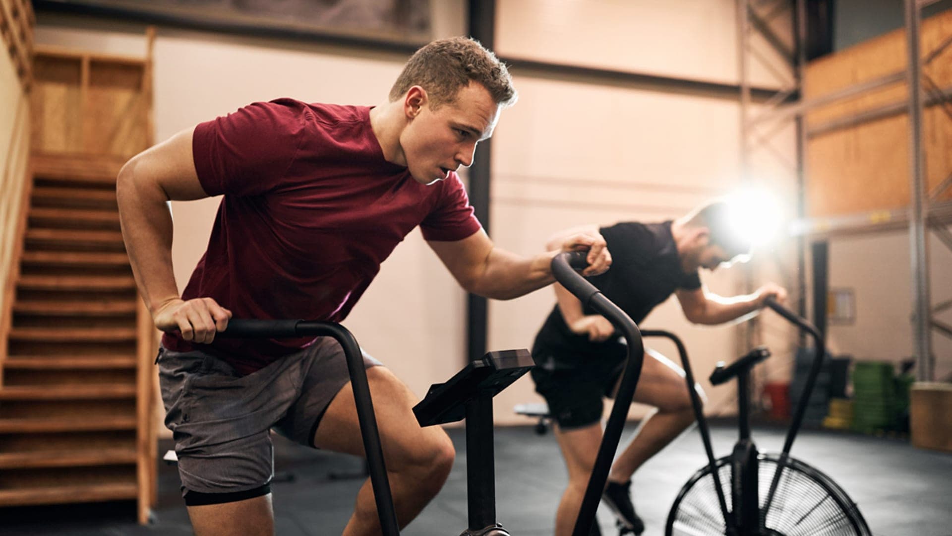 Two men push through a high-intensity workout on stationary air bikes at a gym.