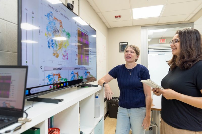 (From left) Kirsten Nielsen and Analis Smith discussing findings from Nielsen's cryptococcus research. On the far right, there is a television displaying data charts on its screen.