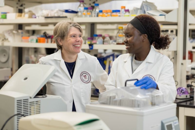 (From left) Kirsten Nielsen and Priscilla Atim working in a laboratory wearing gloves and white coats