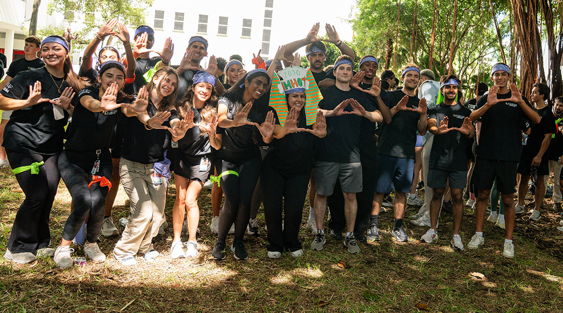 Medical students from the University of Miami Miller School of Medicine Class of 2030 gather outdoors during orientation field day activities. Participants wearing matching black event shirts and colored bandanas stand together beneath trees, forming the University of Miami “U” hand sign while posing as a team.