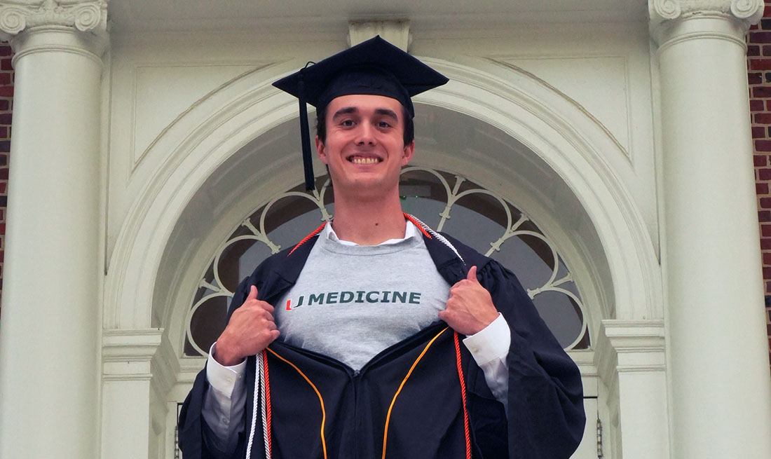 AJ Hernandez, a University of Miami Miller School of Medicine medical student, stands in academic regalia outside a historic campus building with white columns and arched windows. Hernandez opens the graduation gown to reveal a UMedicine shirt beneath.