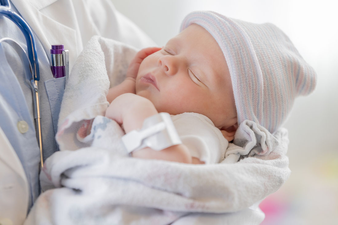 A newborn infant wrapped in a hospital blanket is held by a clinician wearing a white coat and stethoscope, with a hospital ID band visible on the baby’s wrist.