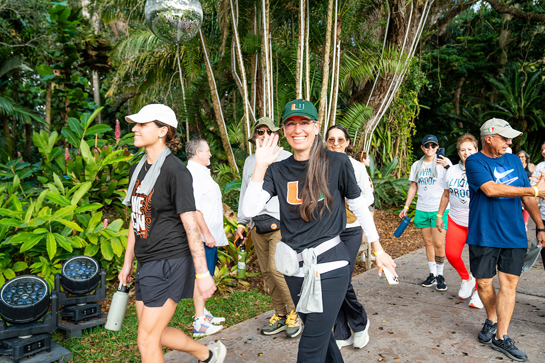 A group of participants walks along an outdoor path surrounded by lush greenery. Several people wear University of Miami apparel, and Donna Scott in the center wears a black UM shirt, green hat, and waves toward the camera. Others walk in both directions, some carrying water bottles or personal items.